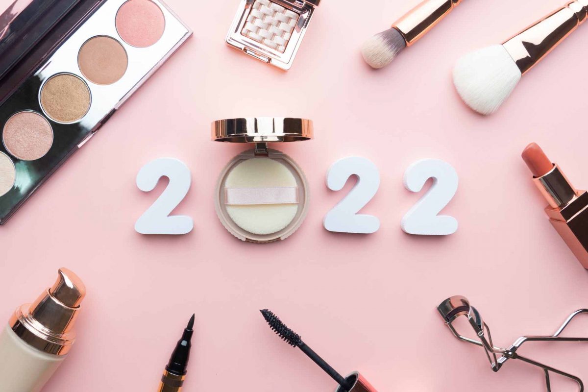 2022 Beauty cosmetic makeup products trends concept. Top view of 2022 white number with powder, lipstick, foundation, eyeshadow, brush, eyeliner and mascara on pink background.
